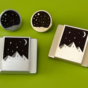 May include: Three silver and gold metal charms featuring a black and white design of mountains, a crescent moon, and stars against a black background.