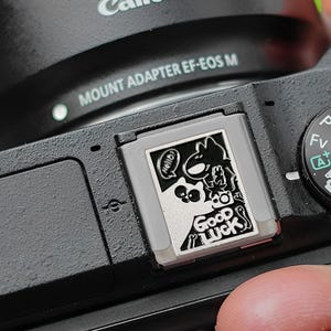 Può includere: Una fotocamera Canon nera con un obiettivo argentato e un adesivo argentato in cima che dice "Buona fortuna".
