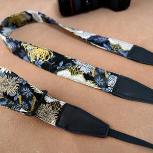May include: A floral camera strap with a black base, featuring yellow, white, and blue flower patterns. The strap has black leather ends and a black adjustable buckle. The strap is designed to be worn around the neck.