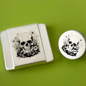 May include: Two silver-toned metal objects with a skull and floral design. One is a rectangular shape, and the other is round. The skull is depicted in black ink, surrounded by flowers and foliage, on a bright green background.