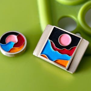 May include: Two pieces of jewellery with vibrant enamel designs. One is a round button with a swirling pattern in black, pink, red, blue, and orange. The other is a rectangular pendant with a landscape design featuring a pink sun, black sky, and colourful mountains.