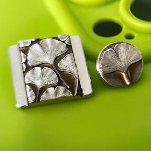 May include: A silver and bronze metal accessory set featuring a ginkgo leaf design. The set includes a square piece and a round button, both with detailed leaf patterns. The items are displayed on a bright green surface.