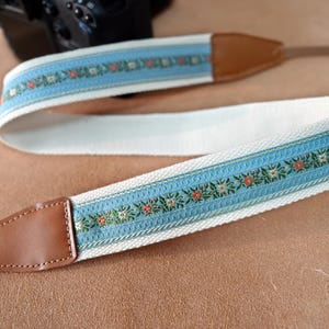 Peut inclure: Une sangle d'appareil photo avec un motif floral bleu clair sur fond blanc. Les extrémités sont en cuir marron. Le motif floral comprend des accents rouges et verts. Conçu pour les appareils photo.