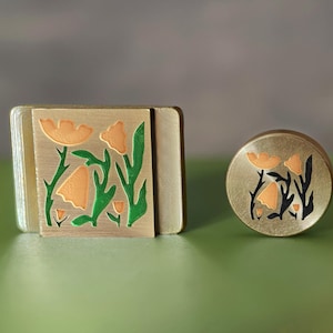 May include: A rectangular brass object and a circular brass object, both featuring an engraved floral pattern with light orange petals and green stems. The metal surfaces have a brushed finish, and the floral designs are recessed into the metal.
