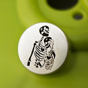 May include: A round, silver-colored metal button featuring a black illustration of two skeletons embracing. The skeletons have heart-shaped details on their rib cages. The button is set against a bright green background.