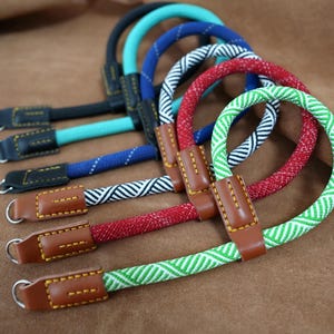 May include: A selection of camera straps in various colours. The straps are in black, teal, blue, red, and green. Each strap has a braided rope design and is attached to a leather connector with a metal ring.