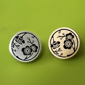 Op de afbeelding: Twee ronde metalen knopen met een bloemmotief. Eén knoop is zilverkleurig met een zwart bloemmotief, de andere goudkleurig met een zwart bloemmotief. De knopen zijn geplaatst tegen een groene achtergrond.