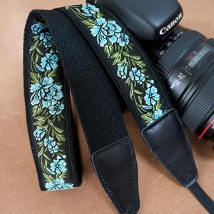 Peut inclure: Sangle d'appareil photo noire avec broderie florale. Le motif répété présente des fleurs bleu clair et des feuilles vertes sur fond noir. Les extrémités en cuir noir et un objectif Canon sont visibles en arrière-plan.