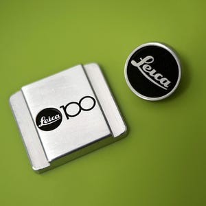 Può includere: Due oggetti metallici su una superficie verde. Uno è un quadrato argentato con il logo Leica e il numero 100 in nero. L'altro è un emblema circolare nero e argento con il logo Leica in bianco.