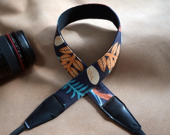 May include: A camera strap with a dark blue background and a repeating pattern of orange and teal leaves and white ovals. The strap has black leather ends and a black interior. A camera lens is visible in the background.