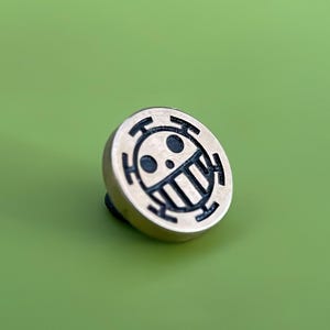 May include: A gold-colored metal pin with a black and white skull and crossbones design. The skull has a toothy grin and a single eye.