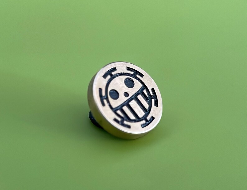 May include: A gold-colored metal pin with a black and white skull and crossbones design. The skull has a toothy grin and a single eye.
