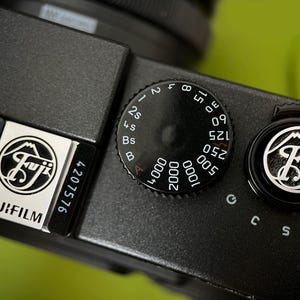 Puede incluir: Primer plano del cuerpo negro de una cámara Fujifilm. La imagen destaca los diales y botones de la cámara, incluido el dial de velocidad de obturación y el interruptor de encendido. El logotipo de Fujifilm es visible.