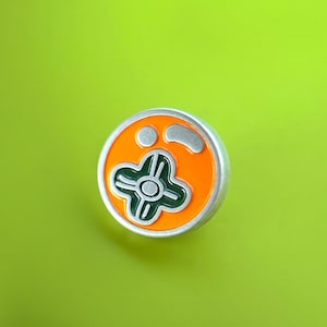 Könnte beinhalten: Runder Ansteckpin mit silbernem Rand und orangefarbenem Innenbereich. Das Design zeigt ein grünes und weißes vierblättriges Kleeblatt in der Mitte, mit zwei kleinen Kreisen und einer gebogenen Form darüber. Vor einem hellgrünen Hintergrund.