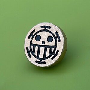 May include: A round, silver-colored pin with a black skull and crossbones design. The skull has a toothy grin and two black eyes.