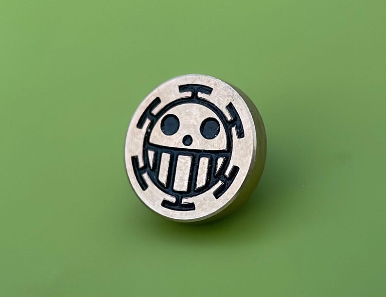 May include: A round, silver-colored pin with a black skull and crossbones design. The skull has a toothy grin and two black eyes.