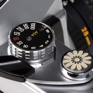 May include: Close-up of a silver camera with a black dial that reads "ASA 1000 500 250 125 60 30 15 8 4 2 1 8 15 30 60 125 250 500 1000". The camera also has a black lever and a white flower-shaped button.