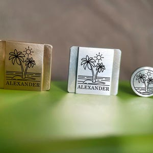 May include: Three metal accessories with a tropical design. Two are rectangular, one gold and one silver, and the third is a silver circle. Each features a palm tree, sun, and the name "ALEXANDER" engraved in black.