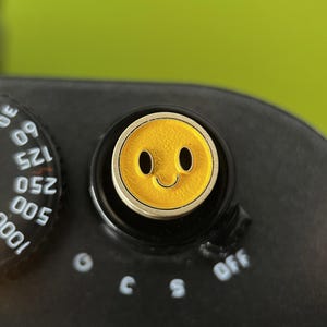 Könnte beinhalten: Eine schwarze Kamera mit einem gelben Smiley-Knopf. Der Knopf hat einen silbernen Rand und ein lächelndes Gesichtsdesign. Die Kamera hat ein Zifferblatt mit weißen Zahlen und Markierungen.