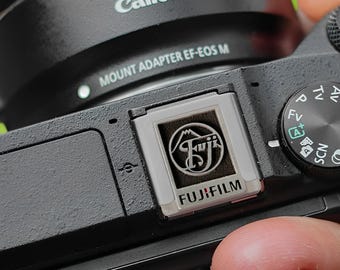 May include: A black camera with a silver Fujifilm logo on the side. The camera has a black lens with the text "MOUNT ADAPTER EF-EOS M" printed on it.