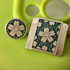 May include: A round and rectangular metal object with a floral design. The design features a light-coloured flower with a dark green background. The objects are on a green surface.