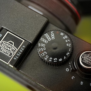 May include: Close-up of a black camera body with a silver dial and the text "NIPPON KOGAKU TOKYO". The dial has numbers from 15 to 4000, and the camera has a silver button with the same text.