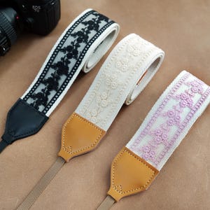 Könnte beinhalten: Drei dekorative Kameragurte mit floralen Mustern. Ein Gurt hat schwarze Blumenspitze auf weißem Stoff, ein anderer cremefarbene Blumenstickerei und der dritte rosa Blumenstickerei. Jeder Gurt hat Leder- und Stoffkomponenten.