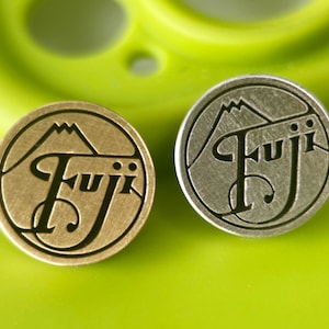 May include: Two circular metal pins, one gold and one silver, displaying a mountain graphic above the word "Fuji" in a decorative font. The pins are set against a blurred green background.