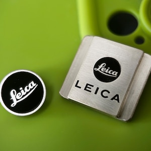May include: Two Leica camera accessories on a green surface. One is a round, black and white emblem with the Leica logo. The other is a silver-coloured, rectangular piece with the Leica logo and text. The background is a blurred green.