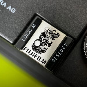 Könnte beinhalten: Nahaufnahme eines schwarzen Kameragehäuses mit einer silbernen Platte mit einem stilisierten Wellendesign und dem Fujifilm-Logo. Die Platte enthält auch den Text "LOICC M" und die Nummer "4207576". Ein Zifferblatt mit weißen Zahlen ist sichtbar.