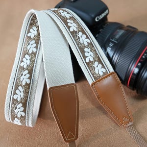 May include: A cream camera strap with a floral design in brown and white. The strap features brown leather ends and is placed on a camera. A Canon camera lens is visible in the background.