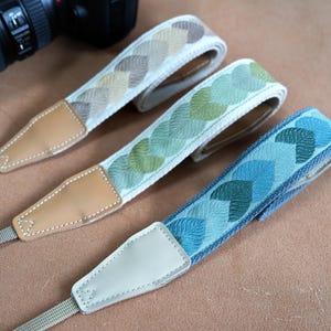 May include: Three camera straps featuring embroidered leaf patterns in beige, green, and blue colorways. Each strap includes tan or light-colored leather end tabs with white stitching and adjustable woven webbing connectors.