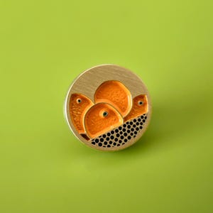 May include: A circular, gold-coloured brooch with an abstract design. It features orange shapes with black dot detailing on a gold background. The brooch is set against a bright green background.