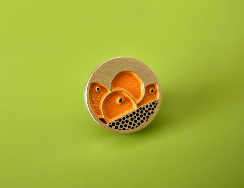 May include: A circular, gold-coloured brooch with an abstract design. It features orange shapes with black dot detailing on a gold background. The brooch is set against a bright green background.