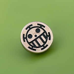 May include: A round, gold-colored metal pin with a black and white design of a skull with a crossbones and a circle around it.