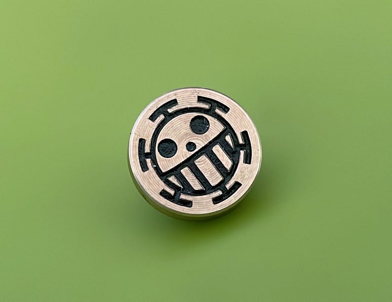 May include: A round, gold-colored metal pin with a black and white design of a skull with a crossbones and a circle around it.
