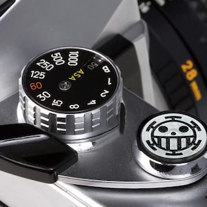 May include: A silver camera with a black dial that reads "ASA" and has numbers from 6 to 1000. The dial is set to 100. There is a black lever on the left side of the camera and a silver button with a black skull and crossbones symbol on the right side.
