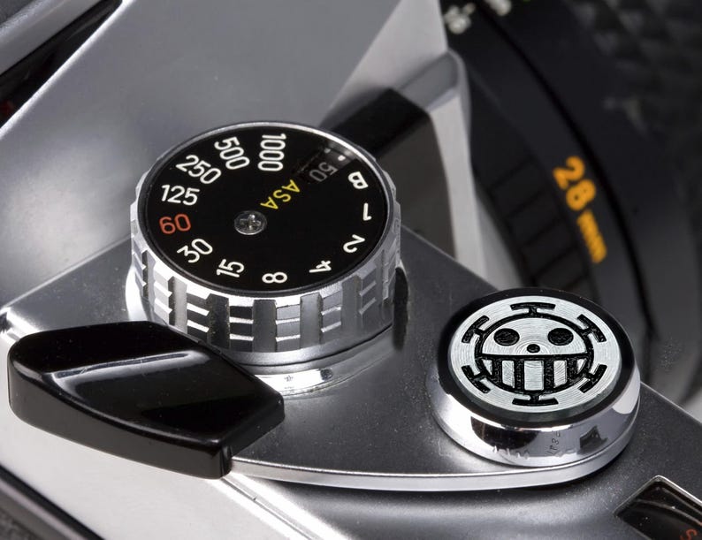 May include: A silver camera with a black dial that reads "ASA" and has numbers from 6 to 1000. The dial is set to 100. There is a black lever on the left side of the camera and a silver button with a black skull and crossbones symbol on the right side.