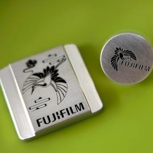 Könnte beinhalten: Zwei silberne Fujifilm Kamera-Zubehörteile. Eines ist eine quadratische Kappe mit einem Kranich-Design und dem Fujifilm-Logo. Das andere ist eine runde Kappe mit dem gleichen Kranich-Design und Fujifilm-Logo. Beide befinden sich auf grünem Hintergrund.