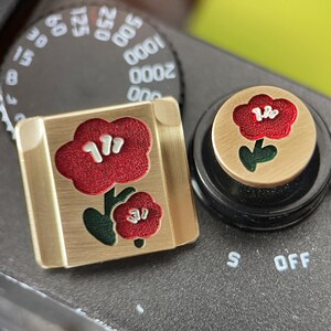 May include: A square and a circular brass-toned metal camera shutter button cover, each featuring a red flower design with green leaves and white accents. The square cover is placed on a camera dial, and the circular cover sits on a black dial.