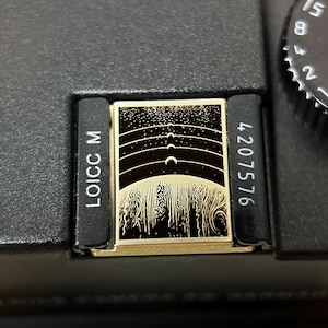 Könnte beinhalten: Nahaufnahme einer schwarzen Kamera mit einer gold-schwarzen Dekorplatte. Die Platte zeigt eine Illustration einer Himmelszene mit dem Text "LOICC M" und der Nummer "4207576".
