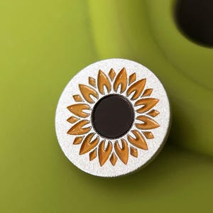 Op de afbeelding: Een ronde, zilverkleurige speld met een zonnebloemontwerp. De zonnebloem heeft oranje bloemblaadjes en een zwart midden. De speld staat tegen een groene achtergrond.