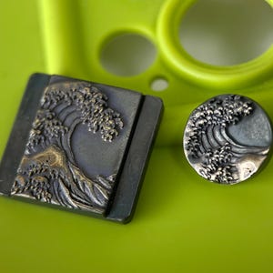 Può includere: Due pezzi di metallo con un disegno a onda. Uno è quadrato, l'altro rotondo. Entrambi hanno una base grigio scuro con un motivo a onda dettagliato in un colore più chiaro, forse oro. Lo sfondo è verde brillante.