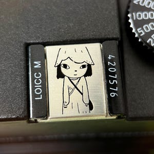 Może przedstawiać: Zbliżenie czarnego aparatu z srebrną płytką przedstawiającą czarno-białą ilustrację postaci w kapeluszu i małej torbie. Na aparacie widoczny jest tekst "LOICC M" i "4207576".