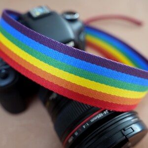 May include: A rainbow-striped camera strap is draped over a black camera body. The strap features bands of red, orange, yellow, green, blue, and purple. The camera lens has the text "24-105mm 1:4 USM" printed on it.