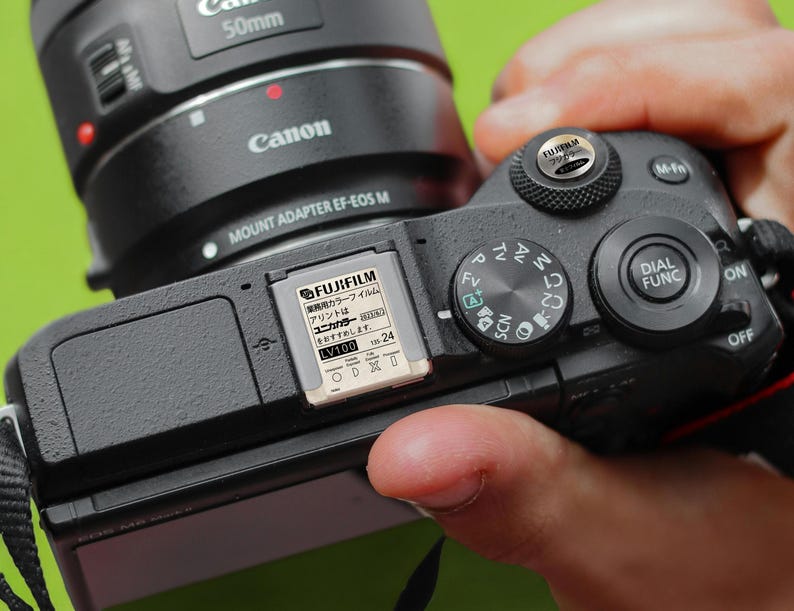 May include: A black Canon camera with a 50mm lens attached. The camera has a black memory card inserted in the card slot. The memory card is labeled "FUJIFILM" and has Japanese text on it. The camera has a variety of buttons and dials, including a dial for selecting the shooting mode, a dial for adjusting the aperture, and a button for activating the flash.