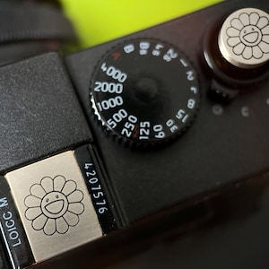 May include: Close-up of a black camera body with a silver dial and a silver button featuring a smiling flower design. The camera has a black dial with white numbers and a silver plate with the text "LOICC M" and a smiling flower design.