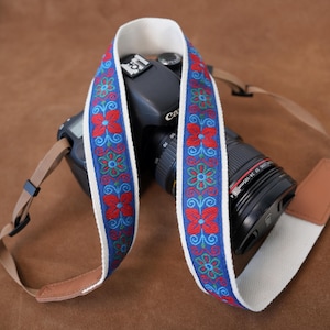 May include: A camera strap featuring a blue fabric centre with red and teal floral embroidery, bordered by white fabric edges. The strap is attached to a black digital camera with a lens, and includes tan leather end tabs with black plastic hardware.