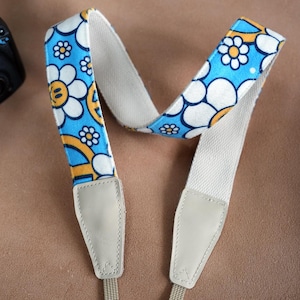 May include: A camera strap with a white background and a blue and yellow floral pattern. The strap has two adjustable leather ends.