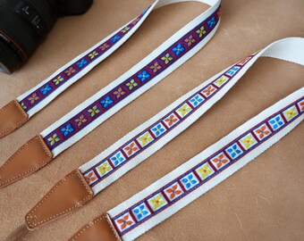 May include: White camera straps with colorful floral patterns in blue, orange, and yellow. The straps have brown leather ends and are designed to attach to a camera. The camera lens is visible in the background.